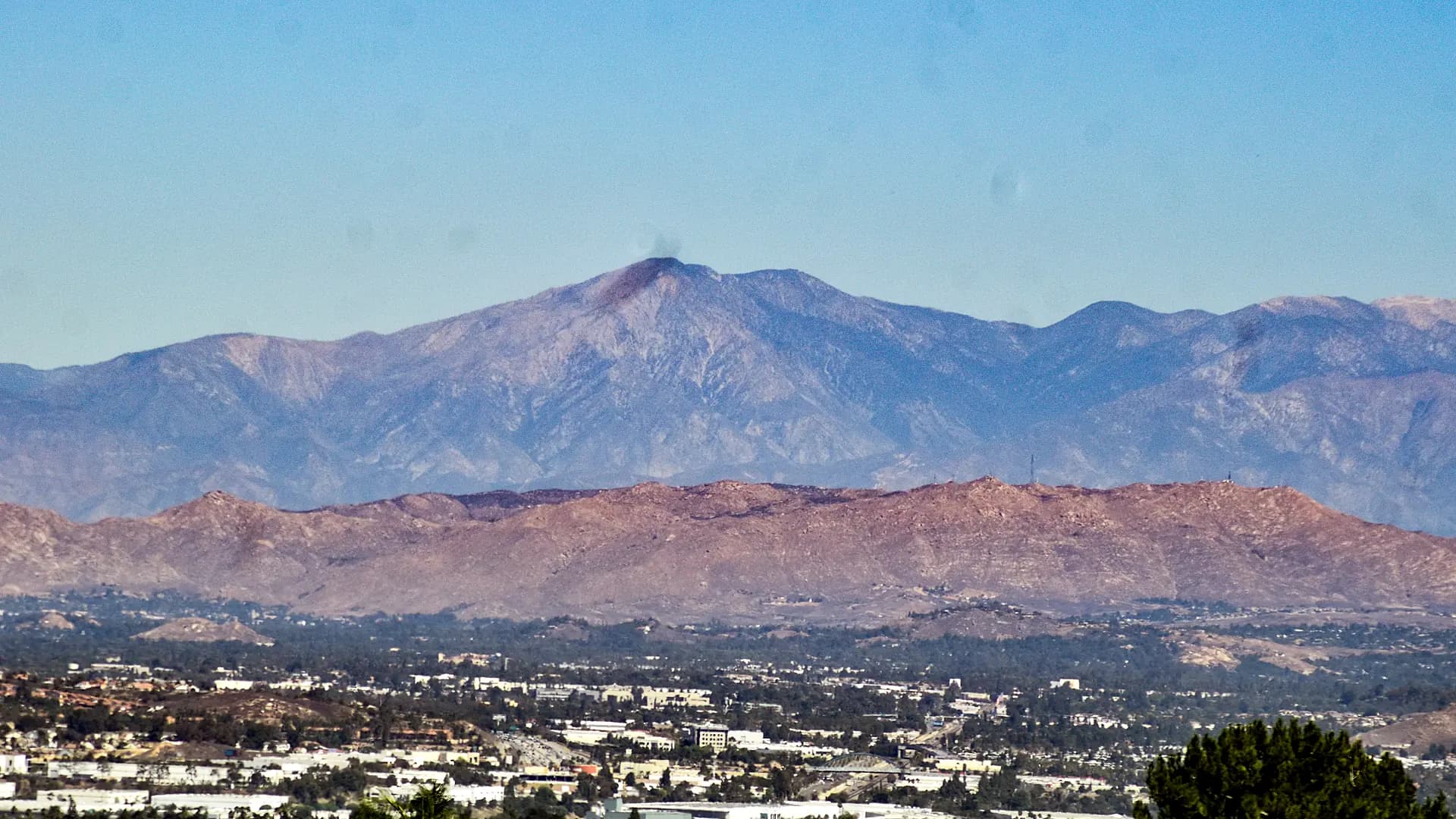 A long view across Corona to the Santa Ana Mountains, the city spread along the valley floor below a red-rock foothill ridge with the higher forested summits behind it