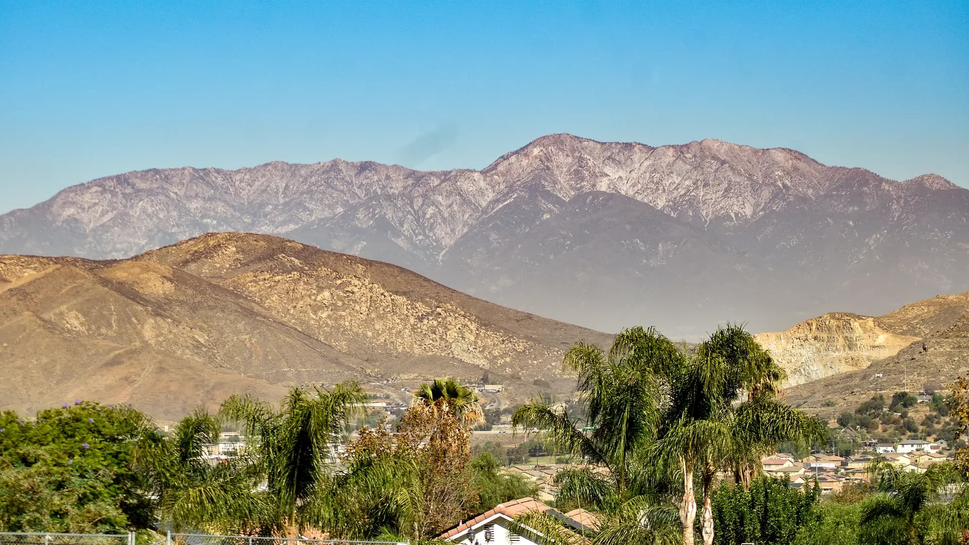 The San Gabriel Mountains rising behind the bare foothills north of Jurupa Valley, queen palms and tile rooftops along the valley floor in the foreground