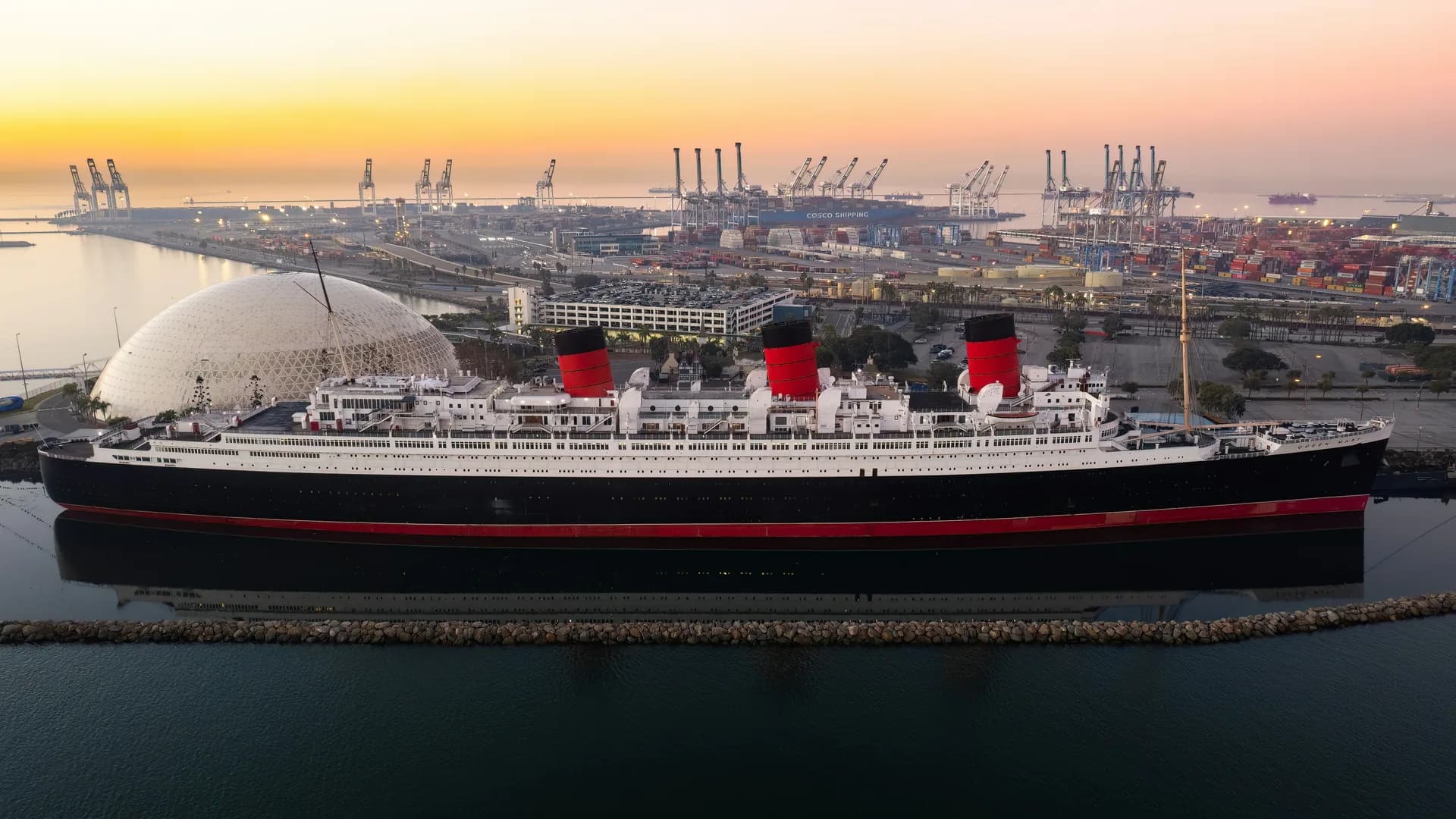 The Queen Mary moored at Long Beach at sunset, the port's container cranes along the shoreline behind her