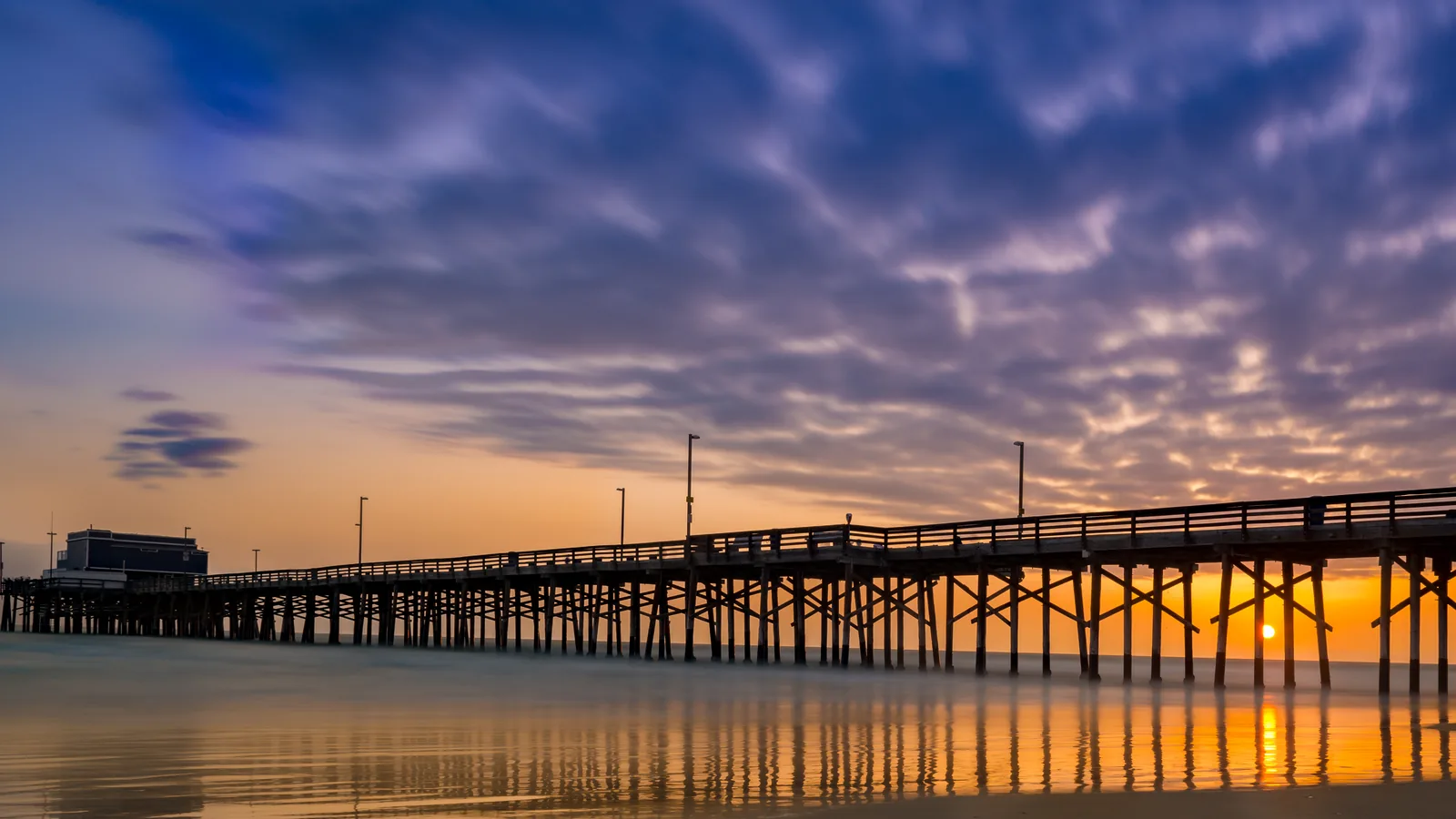 The Newport Beach Pier in silhouette at sunset, its pilings and lamp posts reflected on the wet sand, with the sun on the horizon beneath the deck.