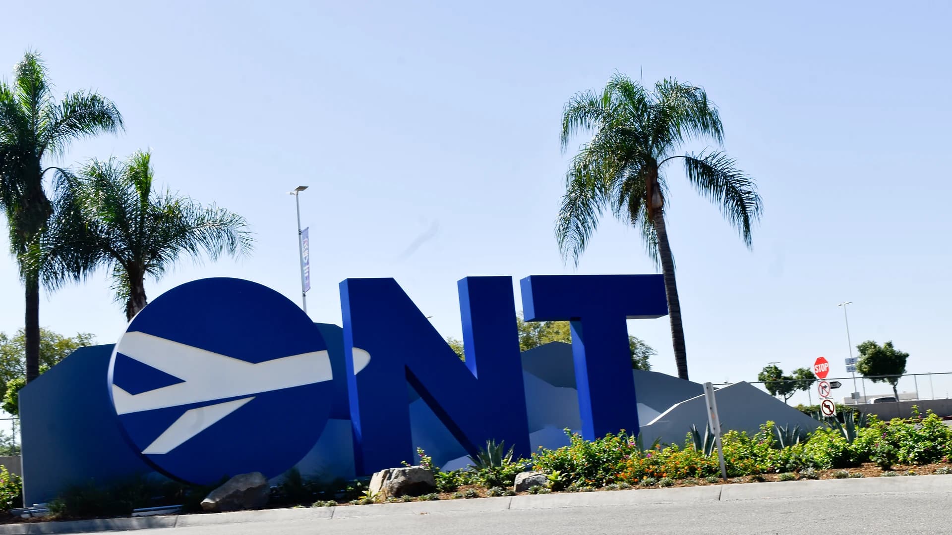 The ONT gateway sculpture at the entrance to Ontario International Airport, oversized blue letters spelling ONT with a jet motif on the O, palms and a clear sky behind