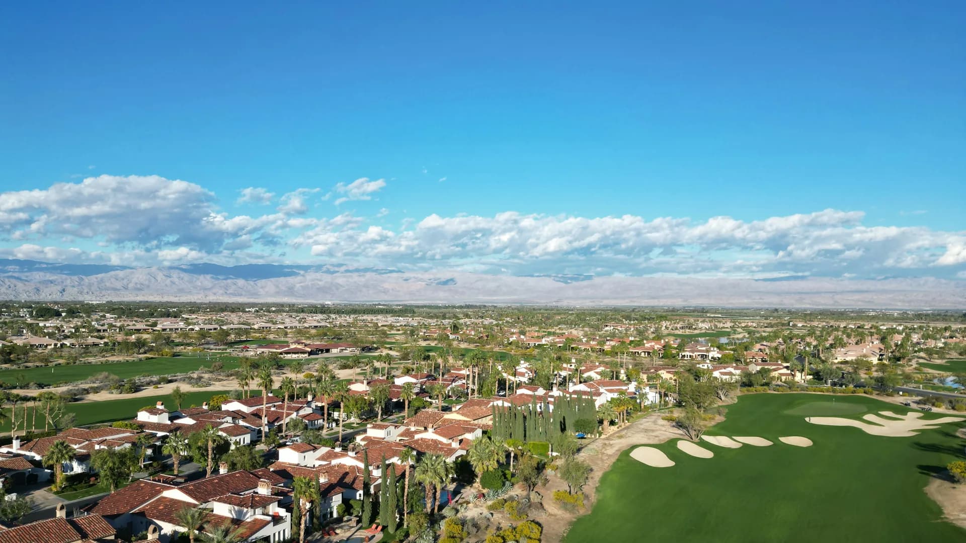 Aerial view over a desert golf course community in the Coachella Valley, low tile-roofed homes among palms with the far side of the valley beyond