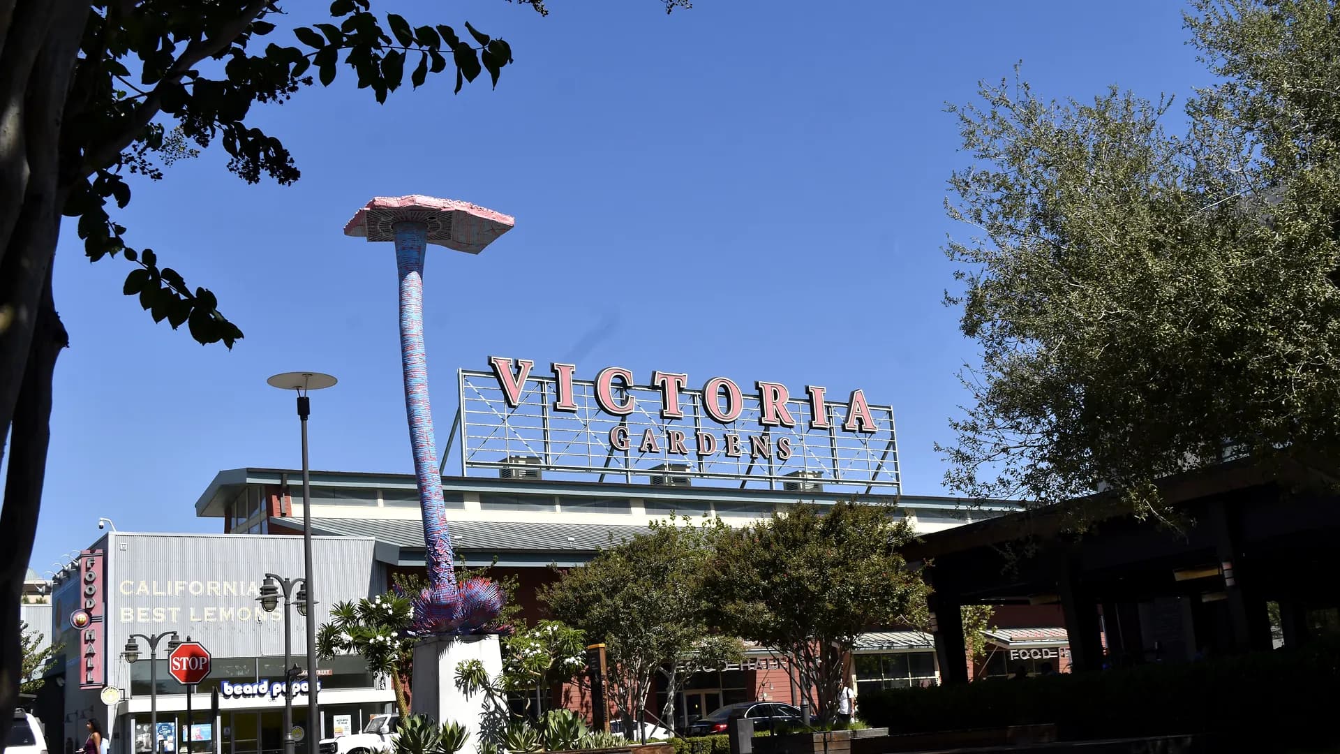The Victoria Gardens sign above the storefronts in Rancho Cucamonga, with the painted mushroom sculpture standing beside it and trees along the street