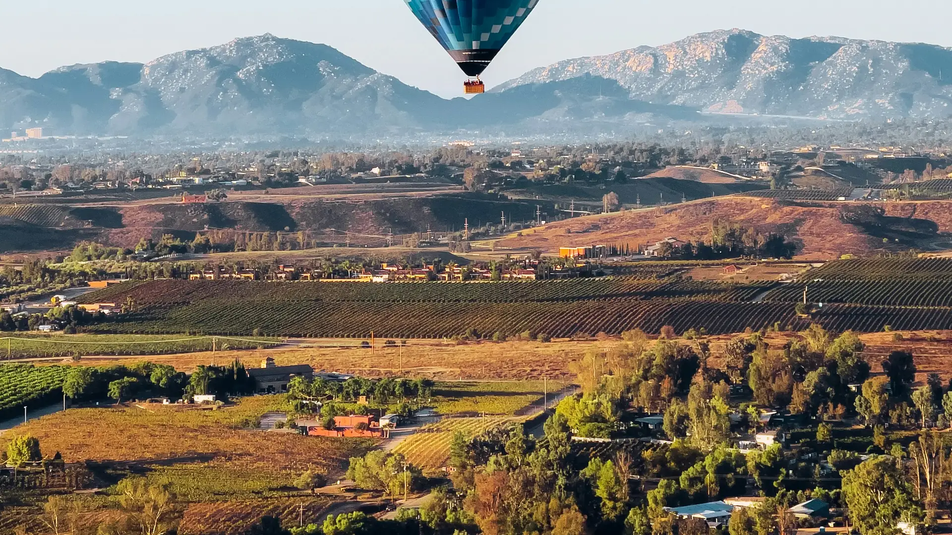 A hot air balloon descending over Temecula Valley at sunrise, vineyard rows across the hillsides and the ridge line behind