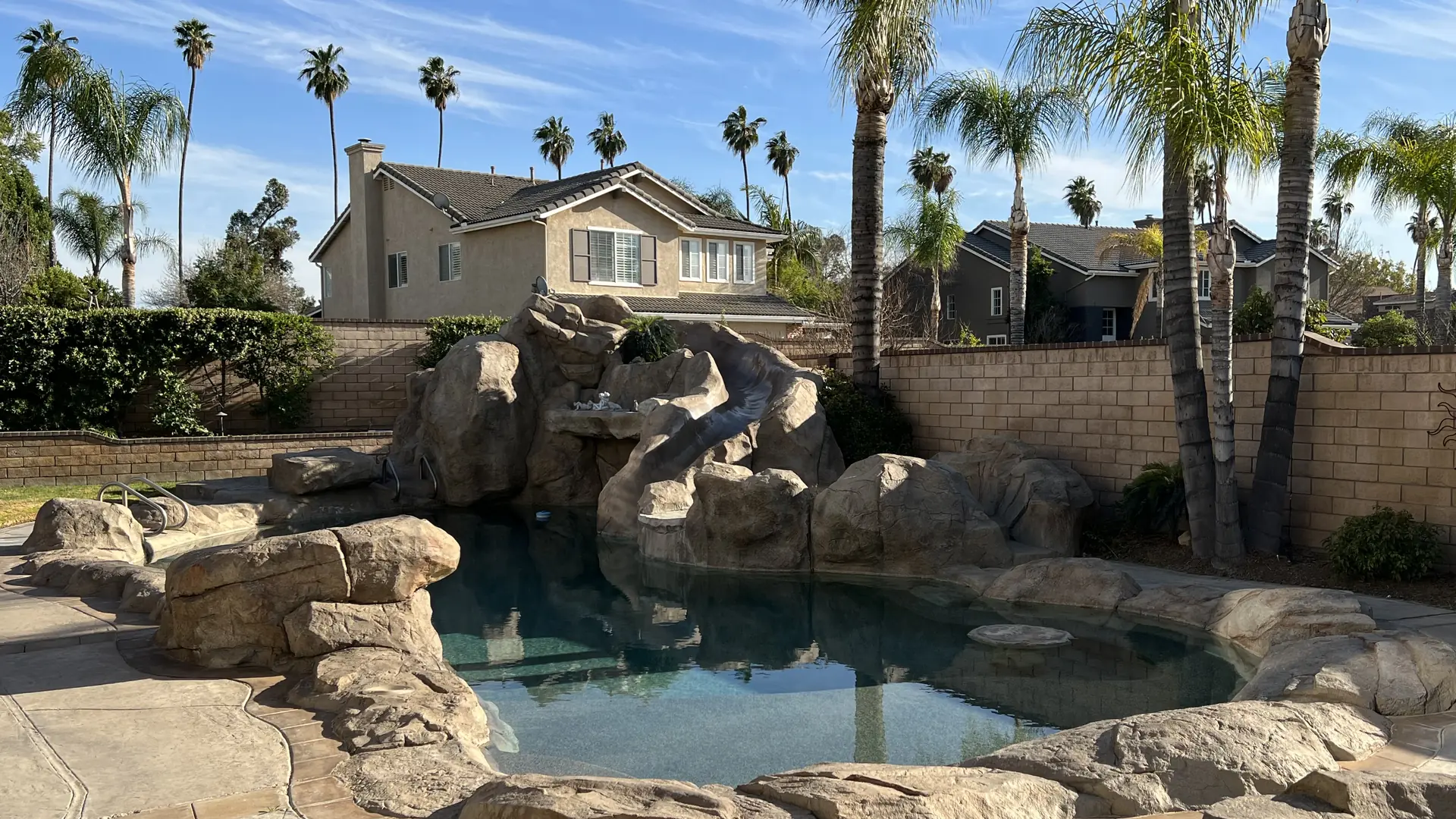 Backyard swimming pool with an artificial rock waterfall and slide, palm trees around it and two-story homes beyond the block wall