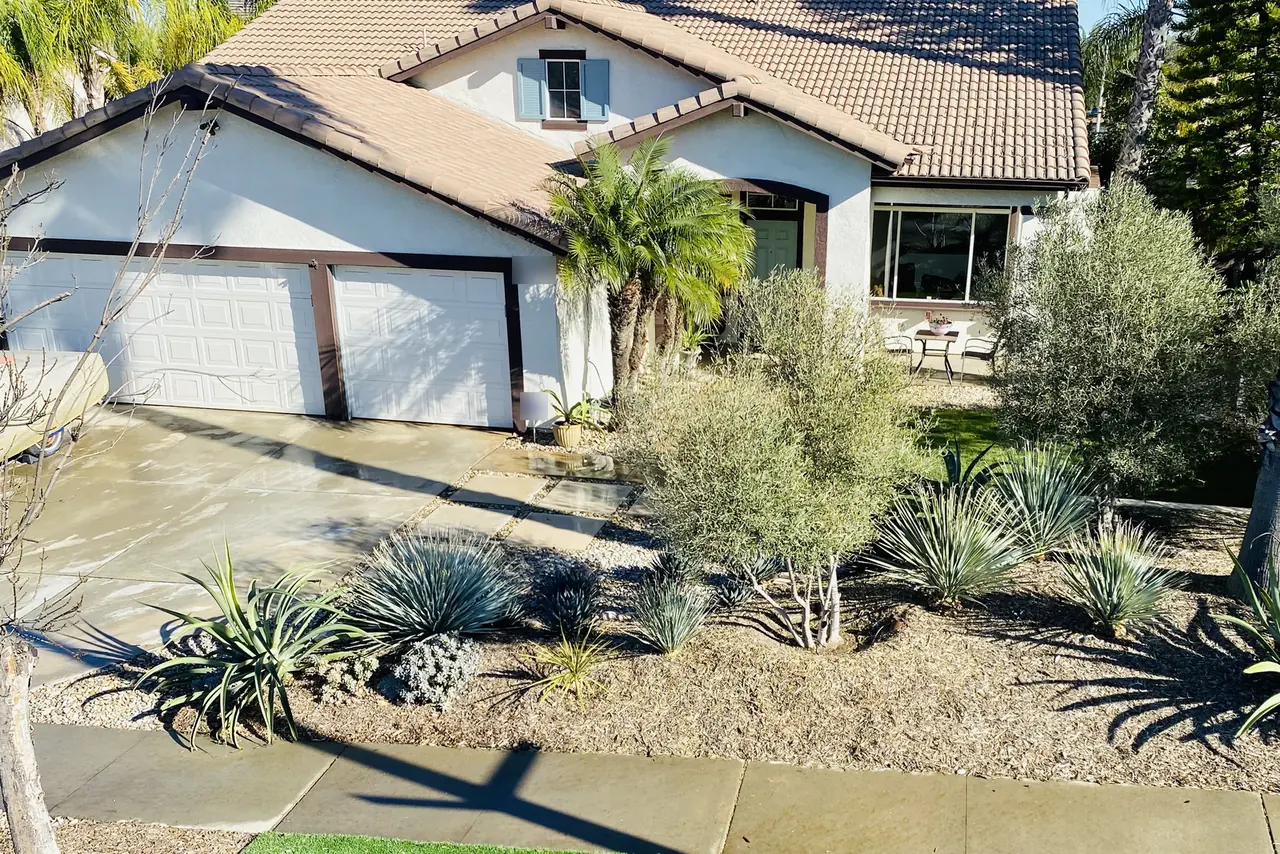 Single-story white stucco home with a tan tile roof and a three-car garage, the front yard planted with agaves, ornamental grasses and an olive tree beside a freshly washed driveway.