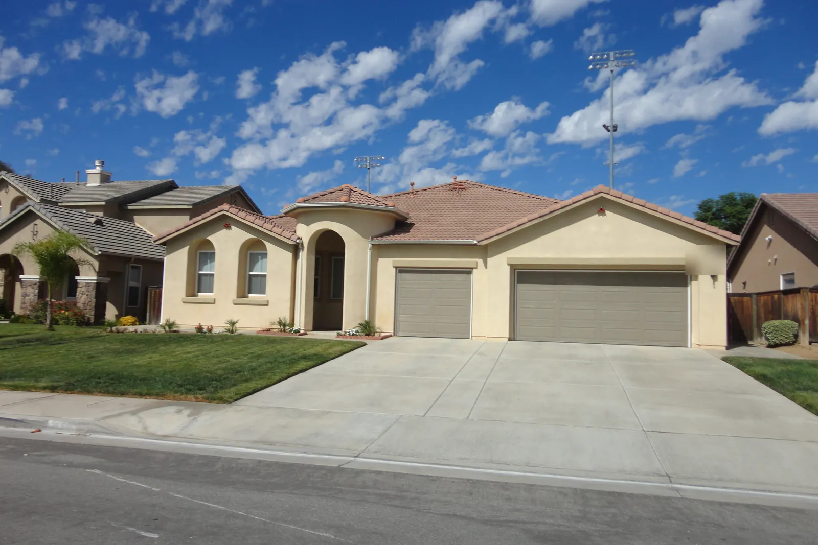 Single-story tan stucco home with a tile roof, an arched entry and two garage doors behind a wide concrete driveway and green lawn, under a blue sky with scattered clouds.