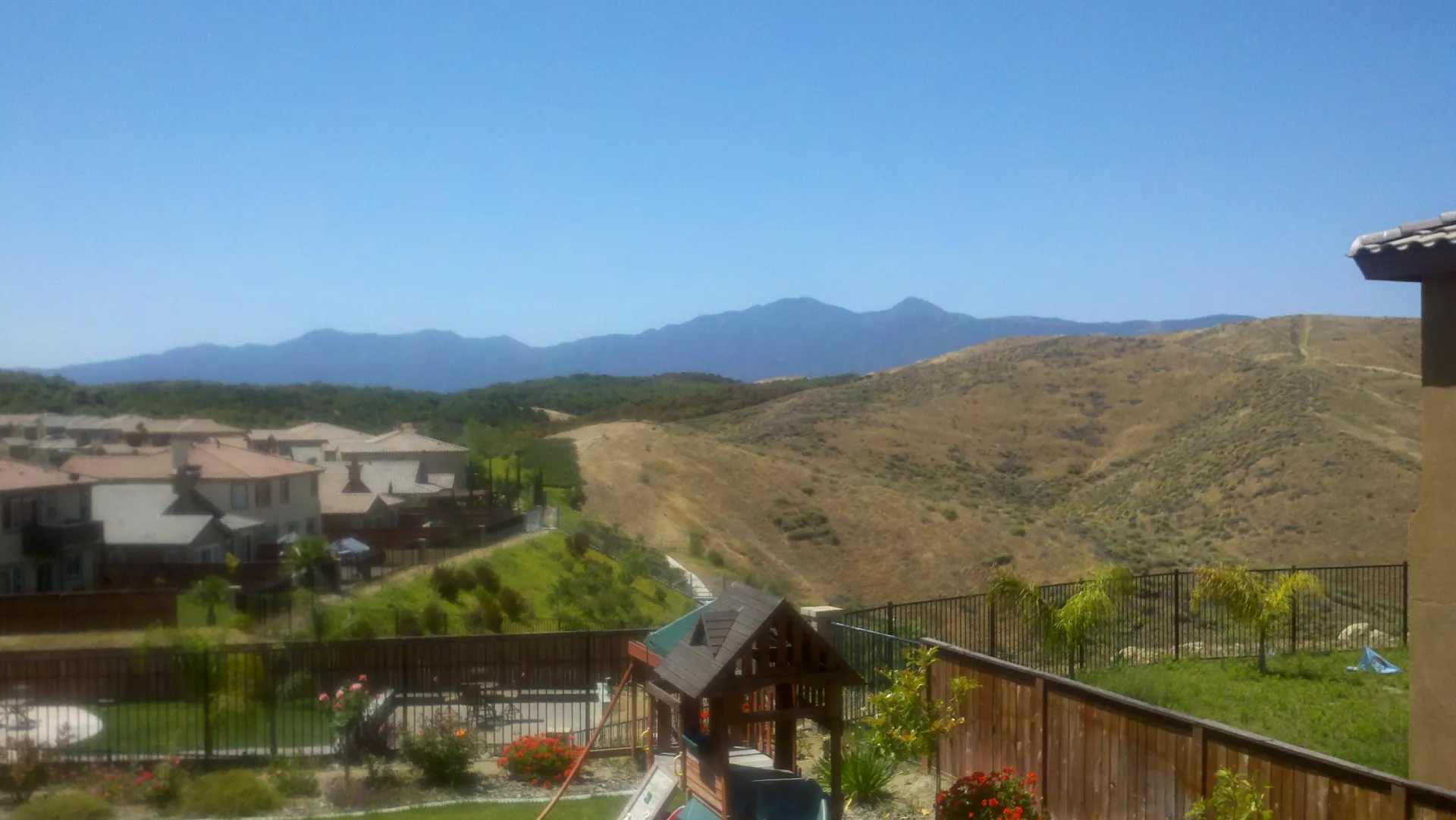 View from a backyard with a wooden play structure and red geraniums, over a row of tile-roof two-story homes and dry golden hills to a blue mountain range under a clear sky.