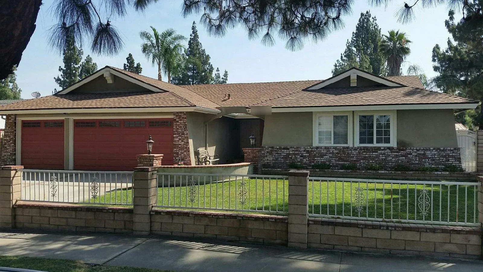 Single-story ranch home with a brown shingle roof, red three-car garage doors and brick trim, behind a block wall with white iron fencing and a green front lawn, with pines and palms overhead.