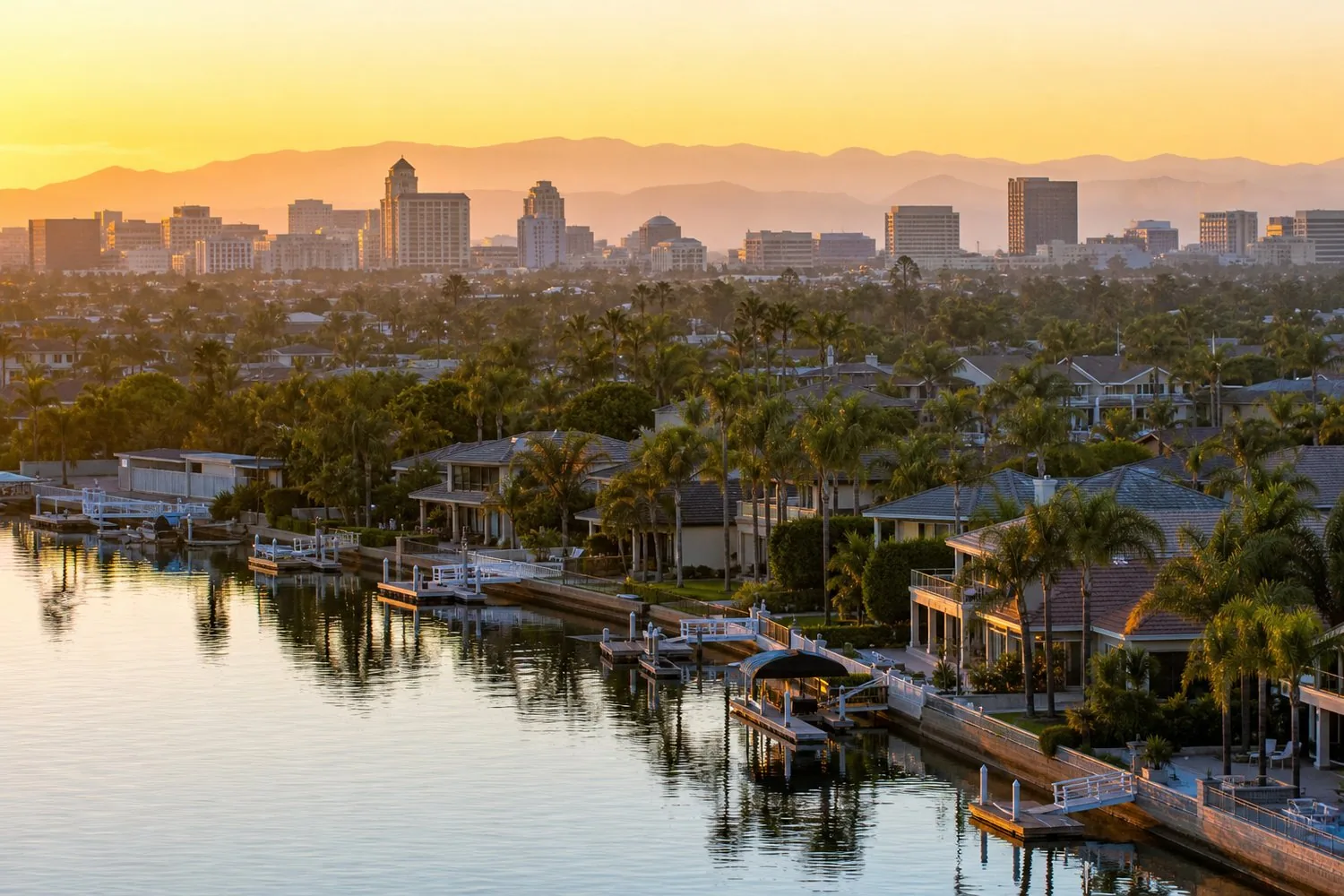 Waterfront homes with private docks along a calm channel at sunset, palm trees and a mid-rise city skyline with mountain ridges behind.