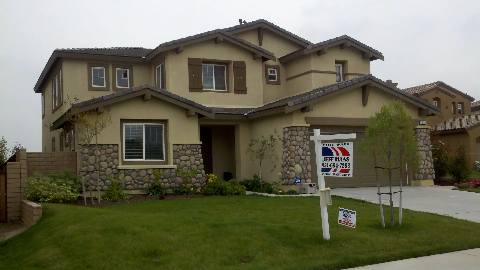 Two-story stucco and river-rock home in a newer Corona subdivision, young street trees still staked along the lawn