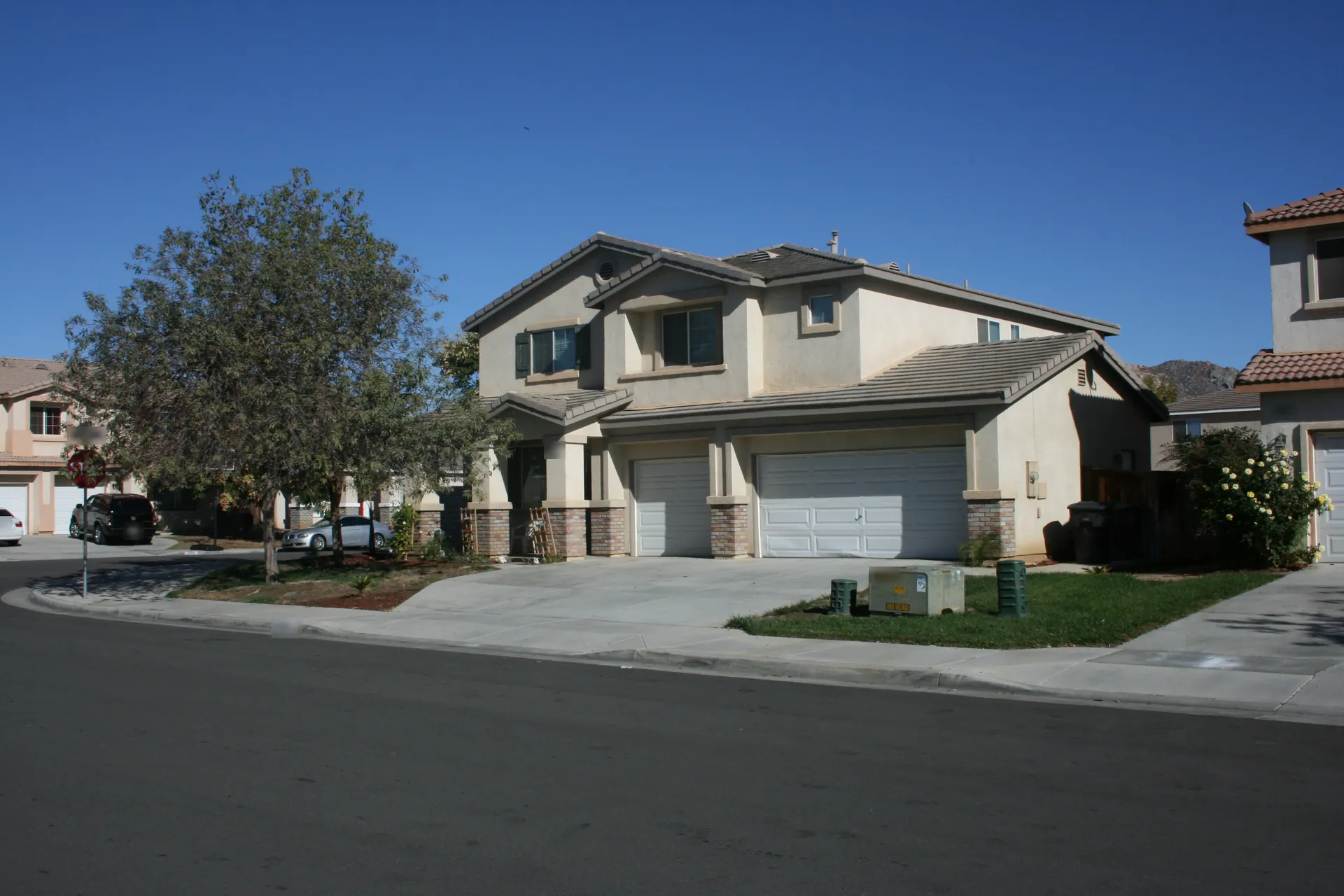 Two-story tan stucco home with a tile roof and three-car garage on a corner lot in Moreno Valley, with a young tree, a green lawn and dry hills under a clear blue sky.