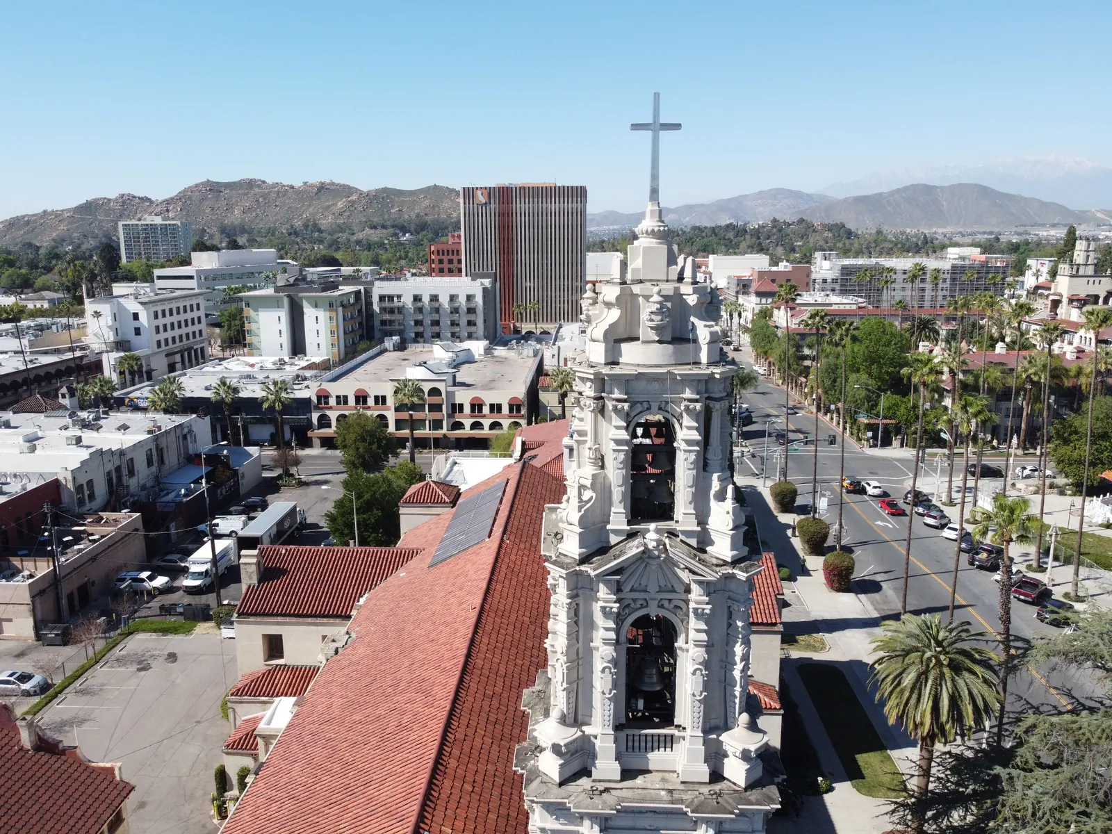 Aerial view over downtown Riverside, California, with the white Spanish Baroque bell tower and red tile roof of a historic church in the foreground, a palm-lined street and low-rise downtown buildings behind it, and the hills beyond.