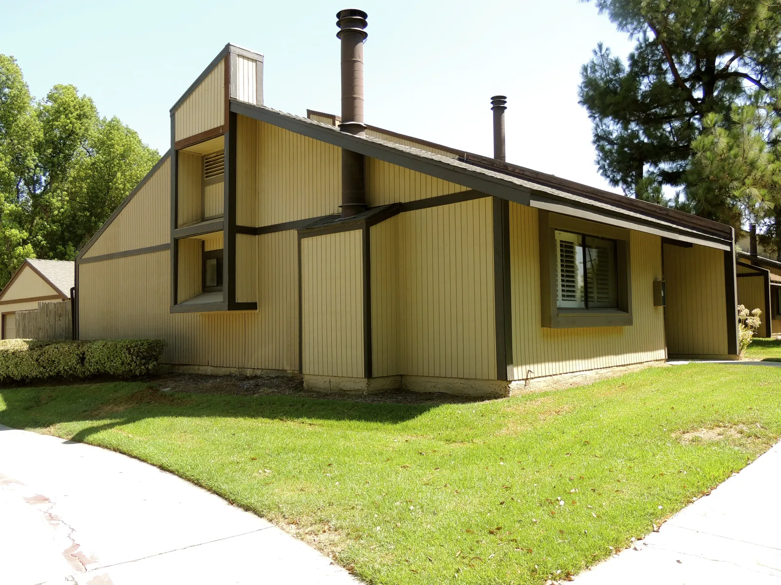 Single-story home with vertical tan wood siding, dark trim and two metal chimney flues on a corner lot with a green lawn and tall pines.