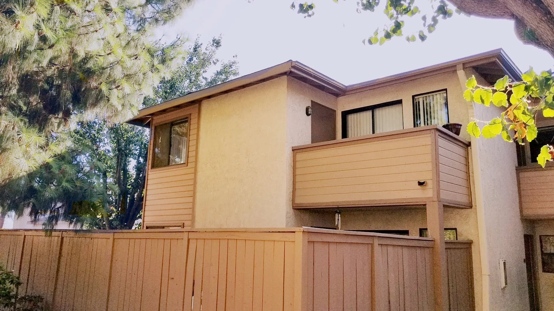 Two-story attached townhouse with an upper-floor balcony behind a wood privacy fence, tree canopy overhead