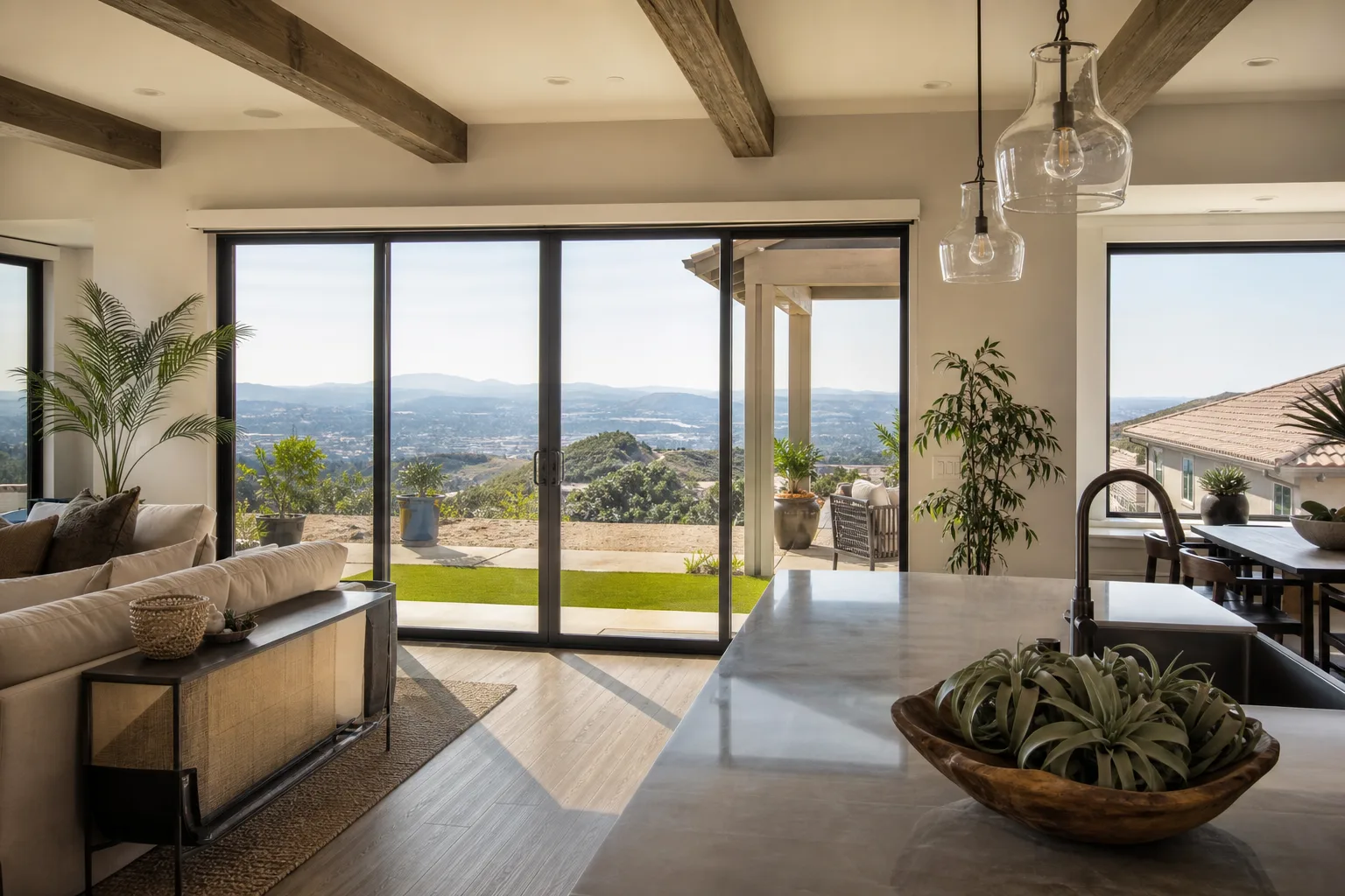 Bright open-plan living room and kitchen with exposed wood ceiling beams, glass pendant lights over a stone island and a wall of sliding glass doors opening to a patio and a wide view of hills and a valley