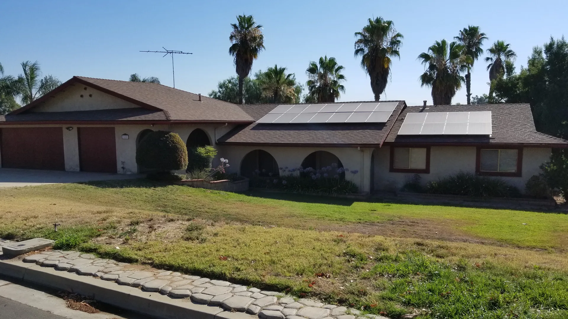 Two roof-mounted solar arrays on a single-story Riverside ranch house, palm trees behind and a stone-edged walkway along the front lawn