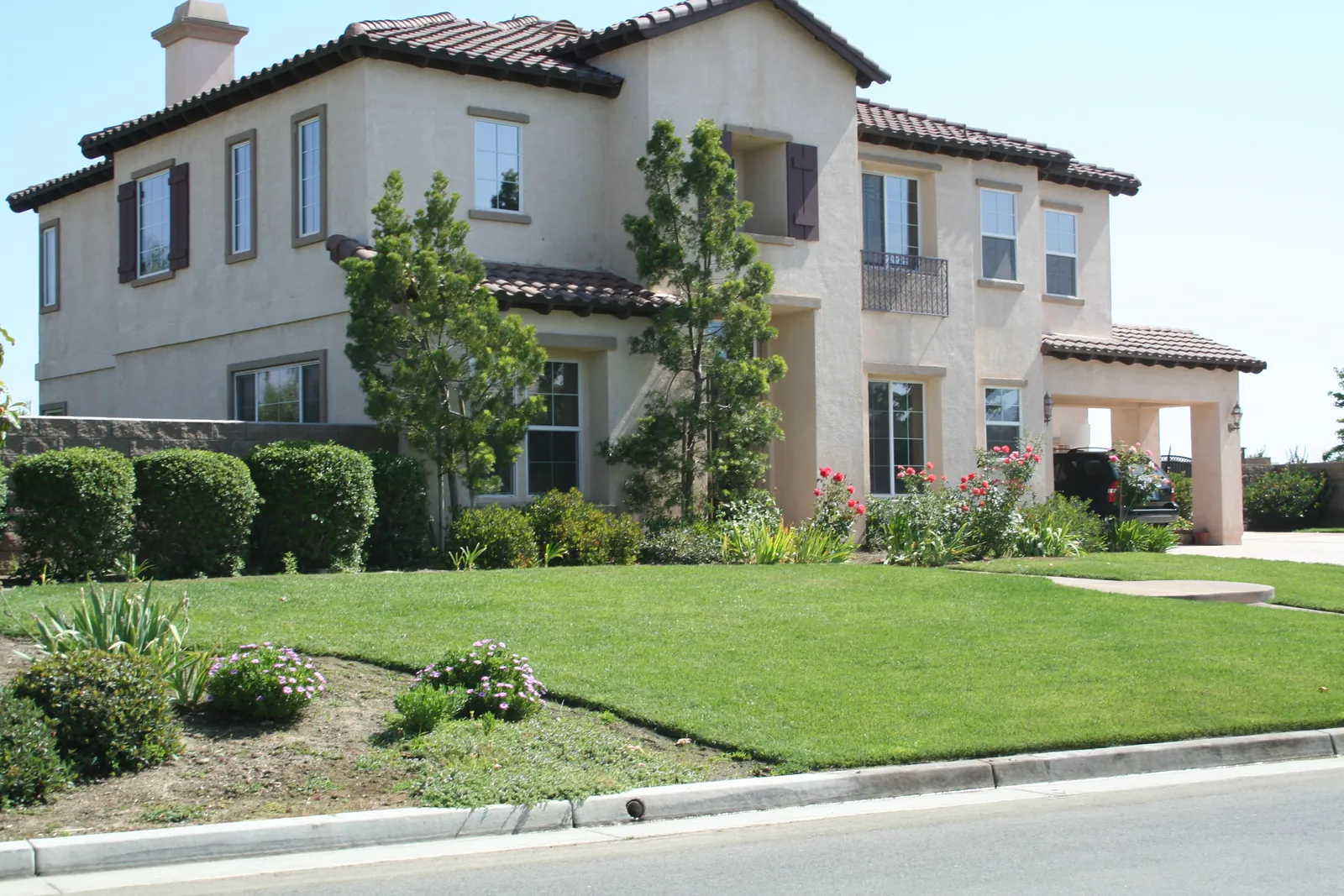 Two-story beige stucco home with dark shutters, a tile roof and a covered carport, set behind a sloping green lawn with rose bushes and young trees.