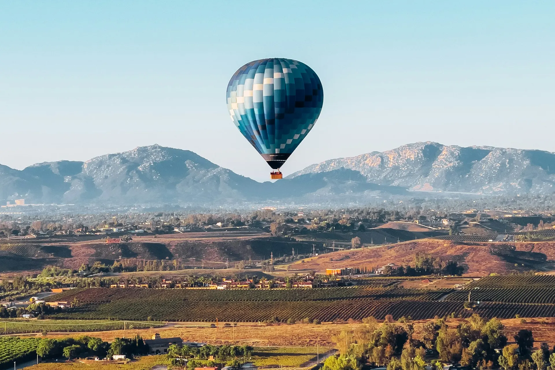 A blue checkered hot air balloon floats over rows of vineyards and rolling hills in the Temecula Valley, with mountains on the horizon under a clear morning sky