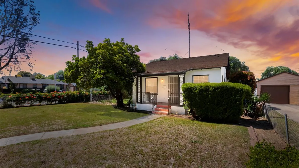 White single-story bungalow photographed at twilight, a mature shade tree on the front lawn and a clipped hedge beside the entry steps